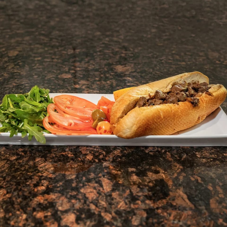 Cheesesteak sandwich with a side of arugula, sliced tomatoes, and pickled peppers on a white plate.