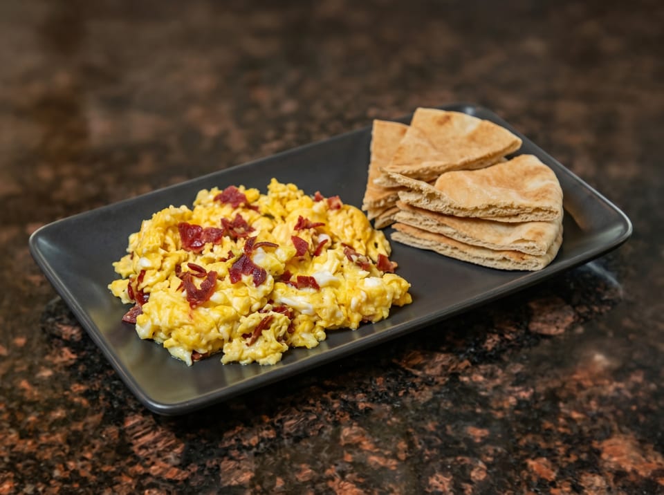 Scrambled eggs with bacon bits and pita bread on a black rectangular plate.