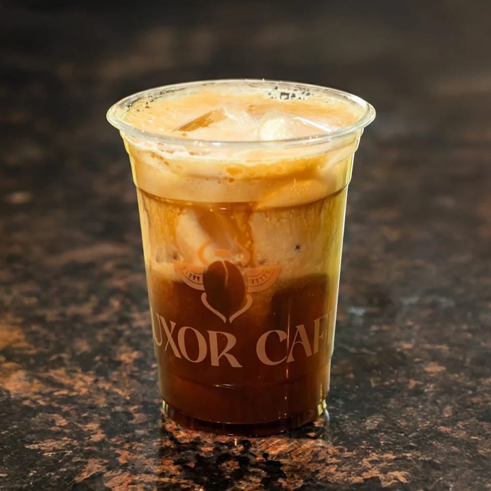 Iced coffee with cream and ice in a clear plastic cup with the Luxor Cafe logo.