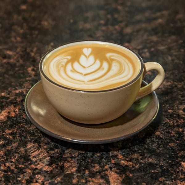 A latte with intricate heart-shaped foam art in a beige cup and saucer on a dark granite surface.