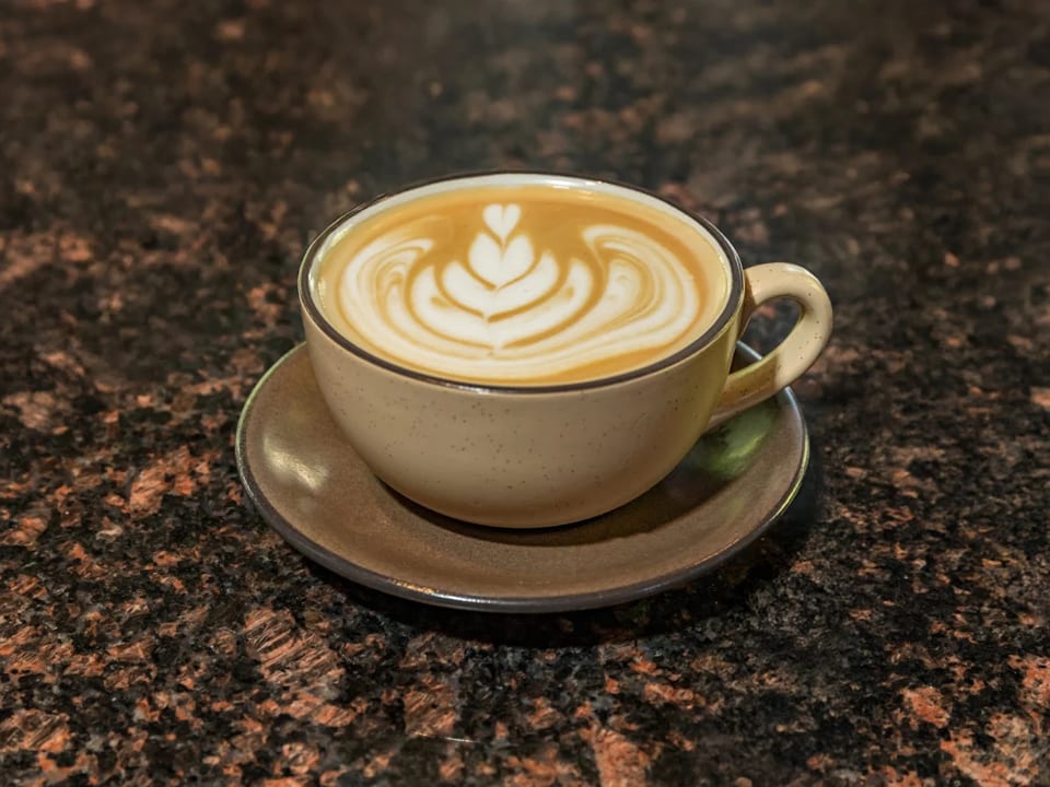 A latte with intricate heart-shaped foam art in a beige cup and saucer on a dark granite surface.