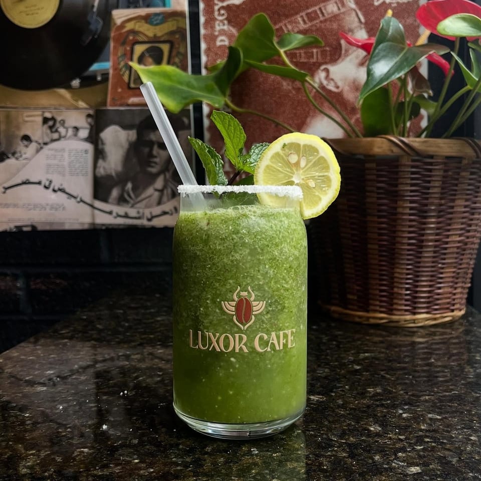 A green Luxor Cafe drink with mint and lemon on a dark counter, with vintage decor in the background.